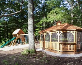 This backyard gazebo has stained wood structure, with screens, and brown shingled roof, and rest on a crushed stone foundation.
