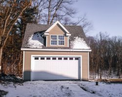 This car garage features tan vinyl siding, white trim, weathered wood shingles, with single dormer.
